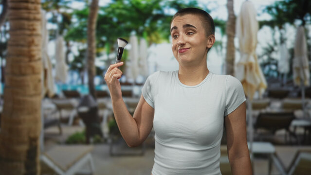 Woman with short hair holds makeup brush up near face with a half shrug expression in building; playful confusion.