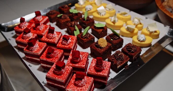 Assorted mini desserts including red velvet cake squares, chocolate brownies, and lemon tarts arranged in rows on a silver buffet tray indoors. Close-up shot of catering pastries at an event.