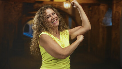 Naklejka premium Woman flexes arm showing biceps and smiles in a studio tunnel corridor set with warm lamps; confidence strength empowerment.