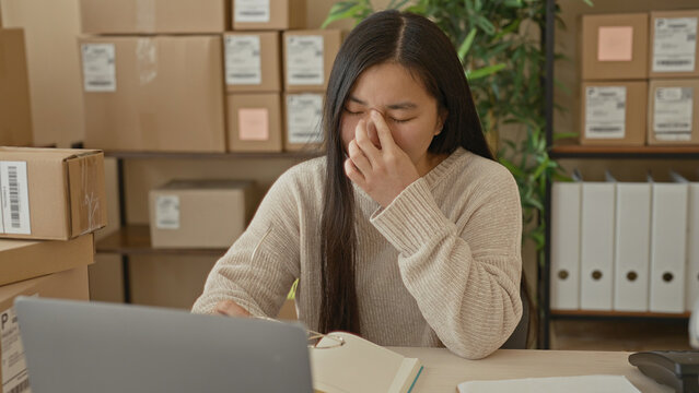 Woman at a packing desk with laptop and cardboard parcels pinches nose and removes glasses while sorting orders in building; work stress.