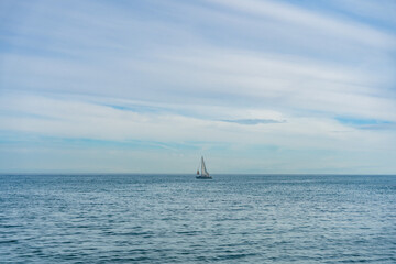 Fototapeta premium Minimal seascape with small sailboat on calm blue ocean under soft cloudy sky. Peaceful marine travel and freedom concept.