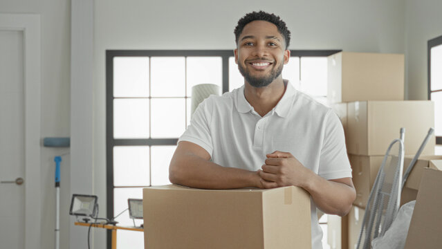 Smiling man holds a cardboard box, rests arm on it and raises fist beside stacked boxes and floodlight in building; moving day confidence.