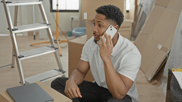 Man holds smartphone to ear while sitting among moving boxes and ladder in a building; focused planning.