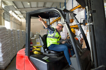 Naklejka premium A warehouse worker navigates a forklift through a warehouse, surrounded by stacked pallets and shelves. He wears a yellow safety vest and a hard hat.