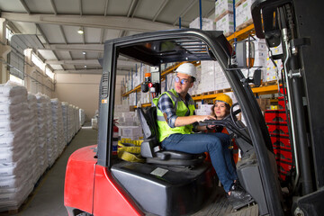 A warehouse worker navigates a forklift through a warehouse, surrounded by stacked pallets and shelves. He wears a yellow safety vest and a hard hat. © bulentbaris