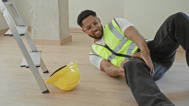 Man clutching injured leg beside fallen hardhat and ladder on a construction floor in a building; accident pain.