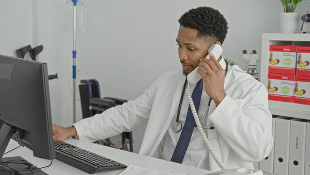 Man holding phone to ear while typing on computer at a clinic desk, stethoscope visible; focused care.