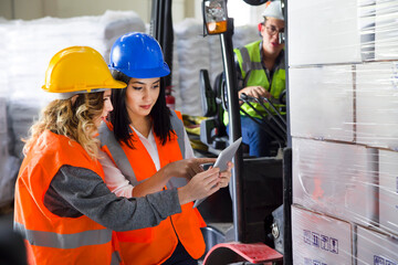 Two women in hard hats and safety vests examine a tablet in a warehouse with a forklift operator working in the background © bulentbaris