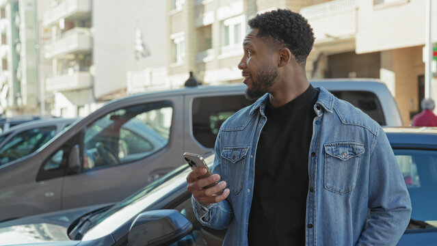 Man holding smartphone in right hand while glancing sideways near parked cars by apartment buildings on a street; thoughtful urban pause.