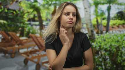 Naklejka premium Young blonde woman with hand on chin beside wooden lounge chairs in forest with tropical plants and trees; pensive contemplation.