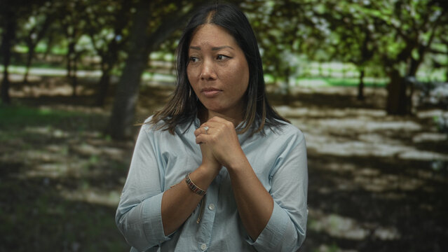Woman clasping hands to chest in park with green trees and dappled sunlight, wearing ring on finger; hope prayer.
