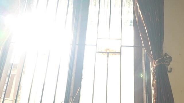 Bright morning sunlight intensely shines through a closed window with dark iron bars, illuminating the sheer white fabric and thick brown side curtains hanging elegantly in the roo