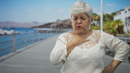Fototapeta premium Woman clutching throat with hand on a seaside boardwalk pier by railing and boats, facial grimace and visible neck gesture showing breathing difficulty; distress.
