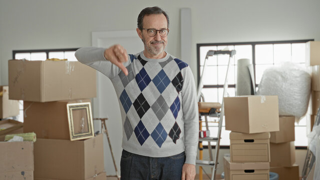 Middle aged man gives thumbs down to stacked cardboard moving boxes, ladder and framed picture in a building during a new home move; frustration.
