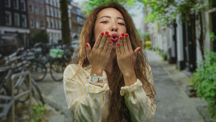 Fototapeta premium Woman blowing a kiss with both hands extended toward camera on a city street lined with bicycles and storefronts; playful affection.