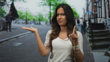 Obraz premium Young hispanic woman holds out hand palm up and points finger on a city street while looking skeptical; skepticism.