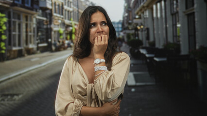 Fototapeta premium Woman biting fingernails with hand to mouth, wearing beige blouse and watch on cobbled city street; anxiety uncertainty.