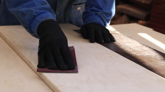 A hand in a black glove sands a wooden surface with sandpaper. The process of manual sanding of a wooden board with fine-grained sandpaper. The craftsman works in a protective glove. DIY concept
