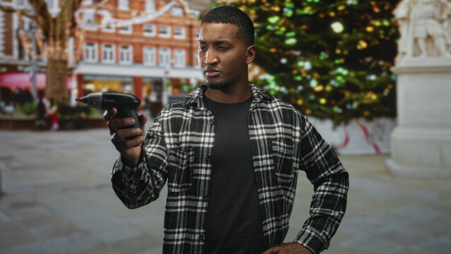Man holding electric screwdriver with right hand, wearing plaid shirt, studying drill tip while hand rests on hip on street near a decorated christmas tree; concentration repair.