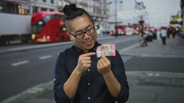 Indonesian man wearing glasses holding a red rupiah banknote between fingers with right hand on busy city street near double decker bus; satisfaction.