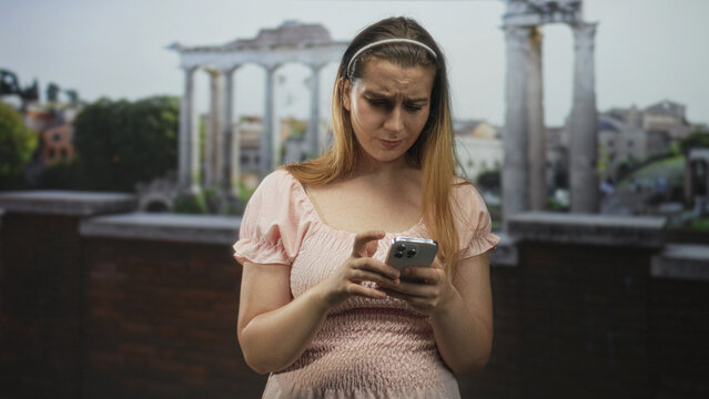 Woman tapping smartphone with furrowed brow in building ruins by ancient columns; frustration uncertainty.