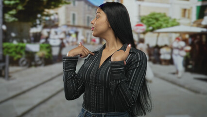 Fototapeta premium Woman in black striped shirt points thumb to chest on sunlit city street with blurred shopfronts and passersby under green trees; pride.