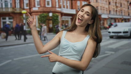 Fototapeta premium Young woman points finger on a city street crosswalk while smiling and gesturing with both hands; playful confidence.