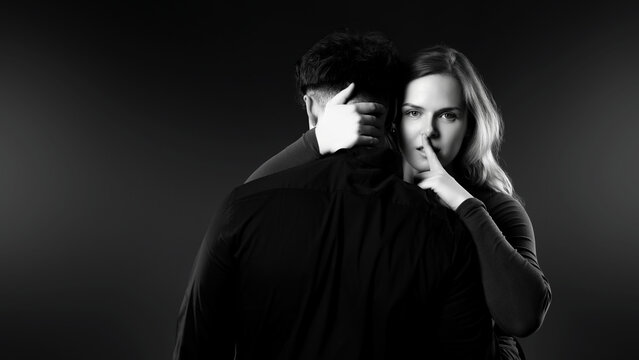 Woman making a shhh gesture to camera while embracing a man, highlighting secrecy