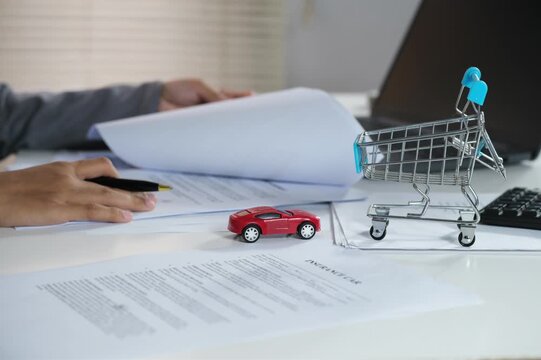An insurance underwriting officer who handles documents, checks documents. Working at a desk with a laptop, pointing to a document with a picture of a small red car on it.
