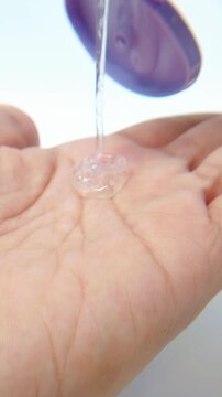 Pouring clear serum onto palm from purple bottle on white background. Close up view of transparent liquid flowing out. Skincare routine, daily beauty treatment.