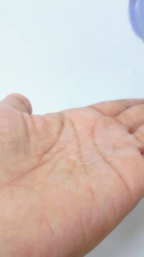 Pouring clear rejuvenating gel serum onto palm from purple bottle on white background. Close up view of transparent serum flowing out. Skincare treatment, anti aging formula.