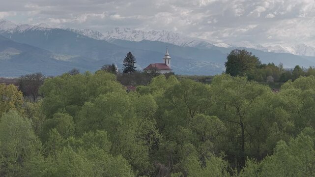 Picturesque white church and swaying green willows in the idyllic rural village of Ucea de Sus against majestic snow capped Fagaras mountains, slow forward flight ungraded log 4K