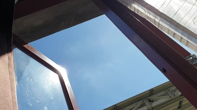 A bright cloudless blue sky is viewed from a low angle right beneath an open wooden window frame, capturing a vivid sun glare on the glass panel alongside a covered white roof area
