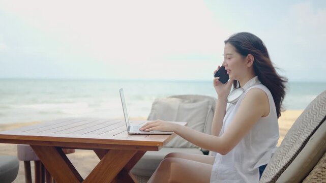 Happy young Asian woman working on laptop and talking on smartphone at beach resort cafe. Smiling female digital nomad enjoying a successful remote work during summer vacation by the tropical sea