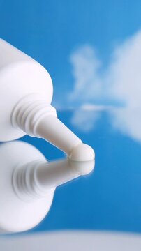 White cream slowly dispensing from white product packaging onto reflective glass surface. Close-up view of thick white cream with soft reflection against blurred blue cloudy background.