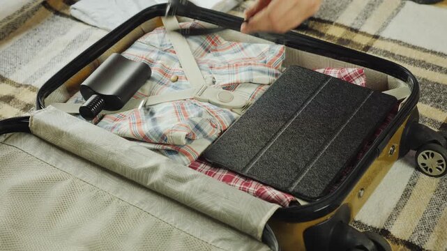 A man carefully placing a tablet, power bank, headphones, and wallet with money into a suitcase before zipping it up.