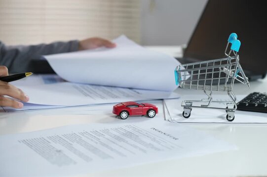 An insurance underwriting officer who handles documents, checks documents. Working at a desk with a laptop, pointing to a document with a picture of a small red car on it.
