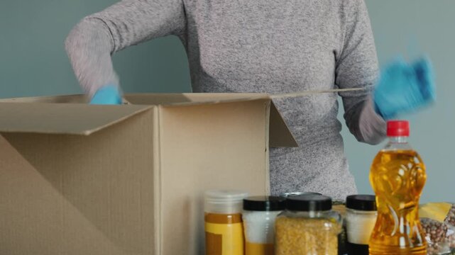 Woman volunteer packing food box for charity, sorting groceries and placing items like oil bottle, spices, and canned goods into cardboard container.
