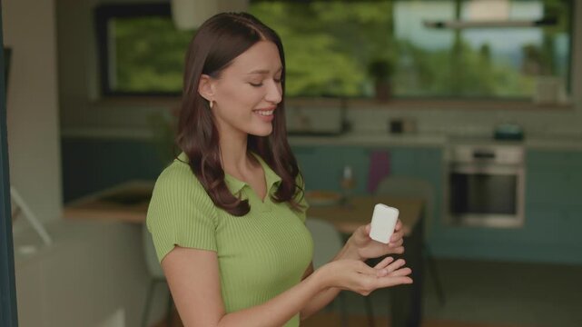 Happy young woman taking vitamin B pill from container at home kitchen, 4K slow motion