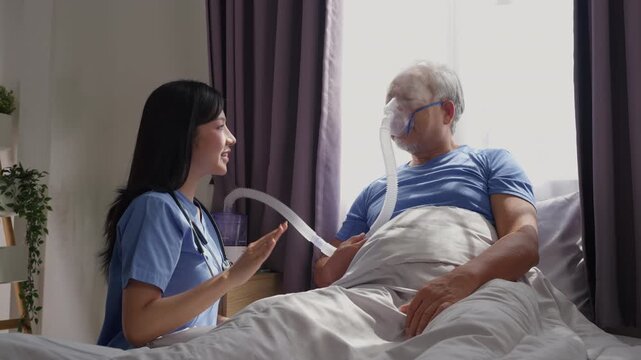 Professional Asian woman caregiver supporting elderly patient man with nebulizer mask breathing treatment oxygen therapy on bed at home. Healthcare Assistance concept.