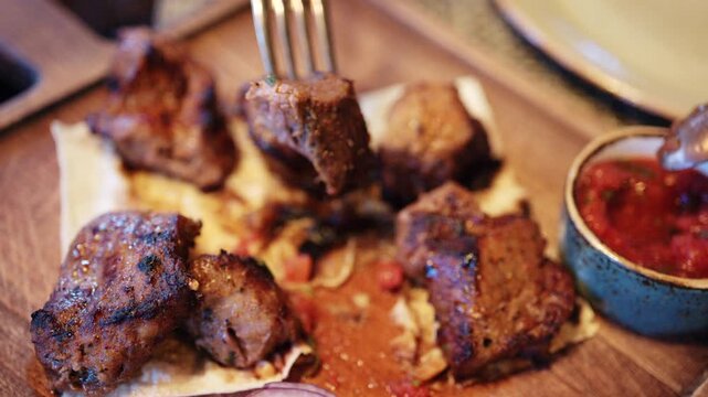 Close up of grilled pieces of meat placed on flatbread, while a person uses a knife and fork to cut into one piece