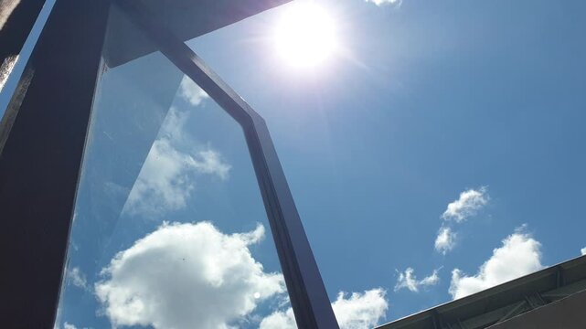 A steep low angle view through an open dark brown window frame captures the glowing sun shining powerfully beside thick fluffy white clouds floating slowly across a pure bright blue