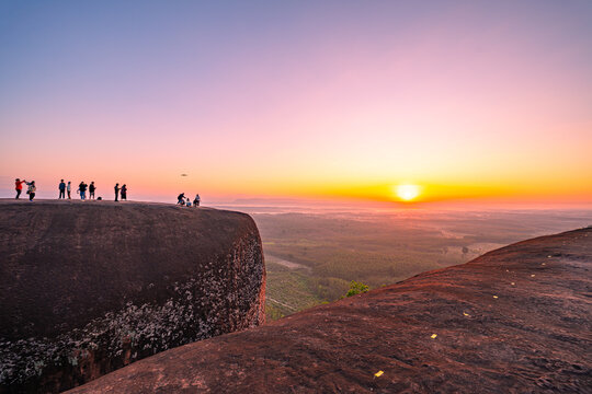 Morning light bathes the sandstone ridge in gold as visitors admire the horizon, their dark silhouettes contrasting beautifully with the expansive natural scenery.