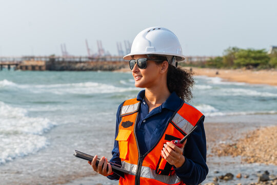 Corporate ESG and CSR industrial compliance: Confident female marine ecologist using tablet and walkie-talkie for offshore terminal logistics and coastal environmental sustainability.