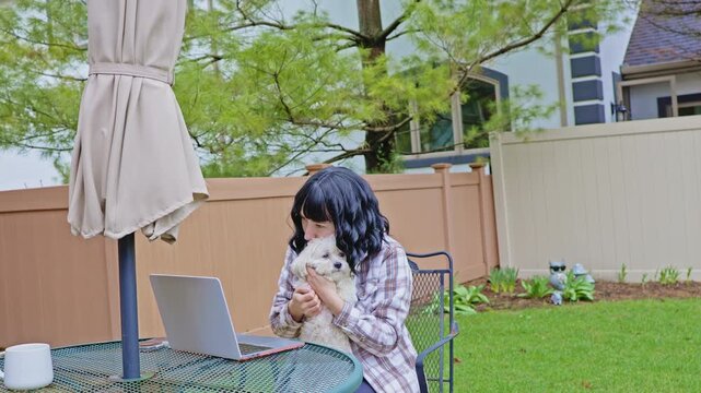 A woman sits at a table in the garden holding a small dog. She works on a laptop while enjoying the warm afternoon. Trees and a fence are in the background.