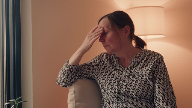 Worried middle-aged woman sitting at home with hand supporting head, deep in thought and emotional distress, 4K