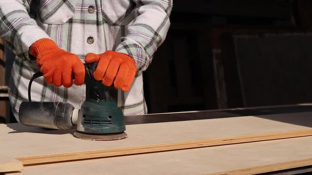 Worker sands wooden surface with random orbital sander. Professional craftsman in protective orange gloves uses orbital sander for finishing plywood. Close-up of woodworking process in workshop