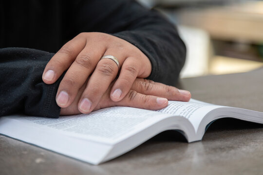 A person's hands are resting on an open religious book. It is daytime, the person is wearing a black sweater and a ring, and they have dark skin.
