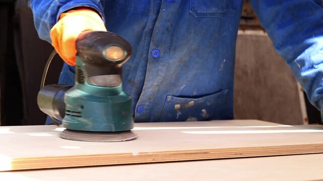 Worker sands wooden surface with random orbital sander. Professional craftsman in protective orange gloves uses orbital sander for finishing plywood. Close-up of woodworking process in workshop