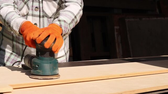 Worker sands wooden surface with random orbital sander. Professional craftsman in protective orange gloves uses orbital sander for finishing plywood. Close-up of woodworking process in workshop
